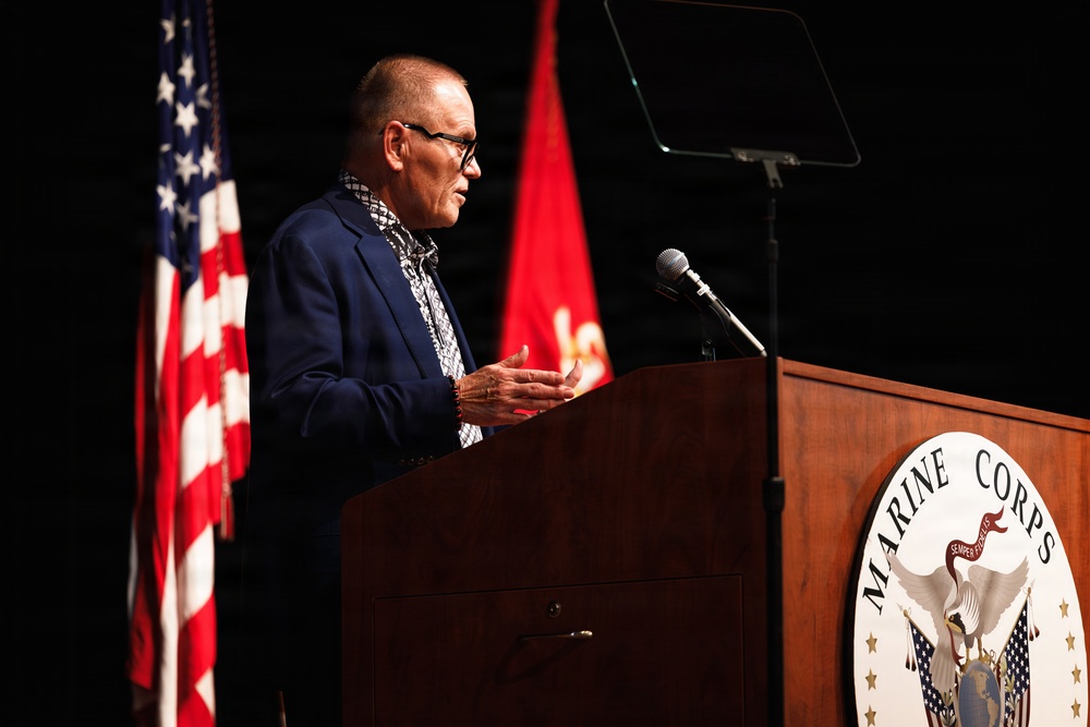 A civilian male speaker addresses an audience from a wooden podium bearing the United States Marine Corps seal. The setting appears to be a formal Marine Corps ceremony or event, flanked by the American flag and a red Marine Corps flag. A music stand or teleprompter is visible behind the speaker.