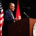 A civilian male speaker addresses an audience from a wooden podium bearing the United States Marine Corps seal. The setting appears to be a formal Marine Corps ceremony or event, flanked by the American flag and a red Marine Corps flag. A music stand or teleprompter is visible behind the speaker.