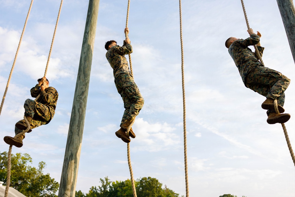 Three individuals in military camouflage uniforms are climbing thick ropes suspended from tall wooden poles against a partly cloudy sky. The scene appears to be a military training exercise or obstacle course, with all three climbers at varying heights and body positions on their respective ropes. The low-angle upward perspective emphasizes the height and physical challenge of the activity.
