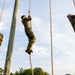 Three individuals in military camouflage uniforms are climbing thick ropes suspended from tall wooden poles against a partly cloudy sky. The scene appears to be a military training exercise or obstacle course, with all three climbers at varying heights and body positions on their respective ropes. The low-angle upward perspective emphasizes the height and physical challenge of the activity.