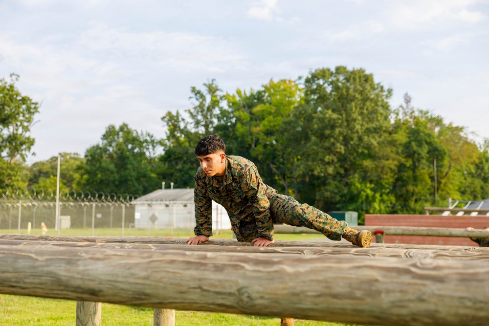 A young military service member in MARPAT (Marine pattern) camouflage uniform is performing a push-up or climbing exercise on a wooden log obstacle. The setting appears to be a military obstacle course or training area, with a chain-link fence topped with razor wire visible in the background. The image is well-lit with natural daylight and lush green trees in the background.