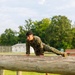 A young military service member in MARPAT (Marine pattern) camouflage uniform is performing a push-up or climbing exercise on a wooden log obstacle. The setting appears to be a military obstacle course or training area, with a chain-link fence topped with razor wire visible in the background. The image is well-lit with natural daylight and lush green trees in the background.
