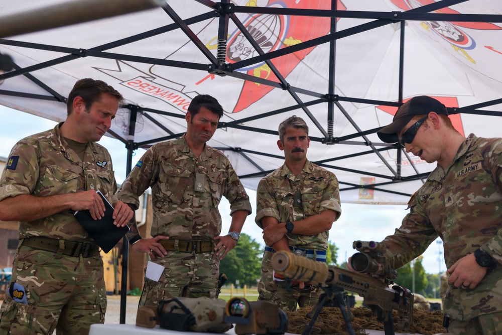 Four military personnel are gathered under a pop-up canopy tent, examining weapons and equipment laid out on a table. Three individuals appear to be wearing British Army MultiTerrain Pattern (MTP) uniforms, while one on the right is clearly identified as U.S. Army by visible tape on his uniform. The setting appears to be an outdoor military demonstration or equipment briefing event.