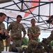 Four military personnel are gathered under a pop-up canopy tent, examining weapons and equipment laid out on a table. Three individuals appear to be wearing British Army MultiTerrain Pattern (MTP) uniforms, while one on the right is clearly identified as U.S. Army by visible tape on his uniform. The setting appears to be an outdoor military demonstration or equipment briefing event.