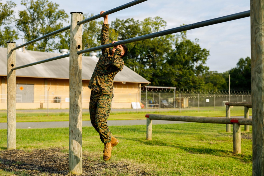 A military service member in full camouflage uniform is navigating a horizontal ladder obstacle on what appears to be a military base obstacle course. The individual is suspended from metal bars supported by wooden posts, demonstrating upper body strength and agility. The setting is an outdoor training facility with manicured grass, fencing, and military-style buildings visible in the background.