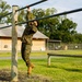 A military service member in full camouflage uniform is navigating a horizontal ladder obstacle on what appears to be a military base obstacle course. The individual is suspended from metal bars supported by wooden posts, demonstrating upper body strength and agility. The setting is an outdoor training facility with manicured grass, fencing, and military-style buildings visible in the background.