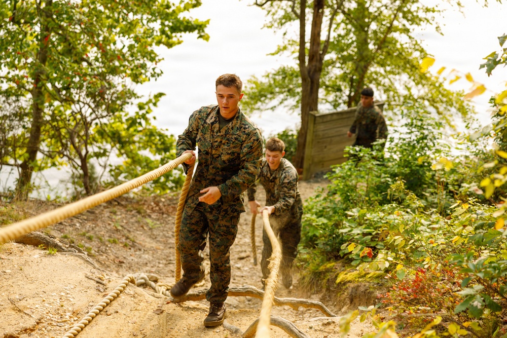 Military personnel in camouflage uniforms are climbing a steep sandy incline using a thick rope for assistance. At least three individuals are visible navigating what appears to be a physical training obstacle course set in a wooded outdoor environment. The scene captures an active training exercise with visible physical exertion.