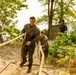 Military personnel in camouflage uniforms are climbing a steep sandy incline using a thick rope for assistance. At least three individuals are visible navigating what appears to be a physical training obstacle course set in a wooded outdoor environment. The scene captures an active training exercise with visible physical exertion.
