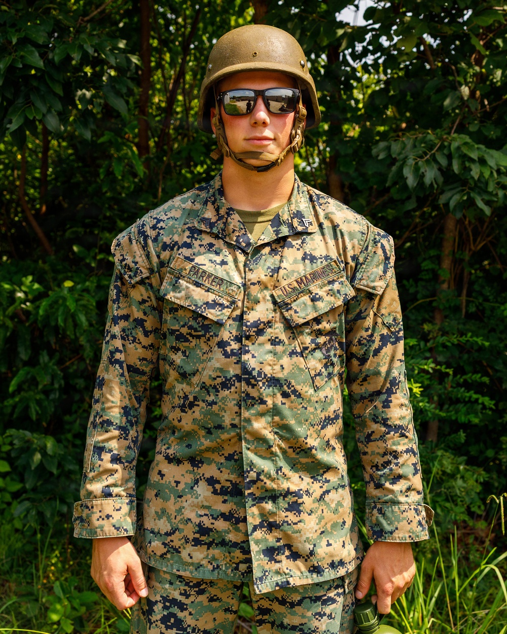 A U.S. Marine identified by name tape as 'PARKER' stands at attention in a wooded outdoor setting. The individual is wearing MARPAT woodland digital camouflage uniform with a Kevlar helmet and reflective sunglasses. The photo appears to be a formal or documentary-style portrait taken in a field environment.
