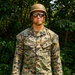 A U.S. Marine identified by name tape as 'PARKER' stands at attention in a wooded outdoor setting. The individual is wearing MARPAT woodland digital camouflage uniform with a Kevlar helmet and reflective sunglasses. The photo appears to be a formal or documentary-style portrait taken in a field environment.