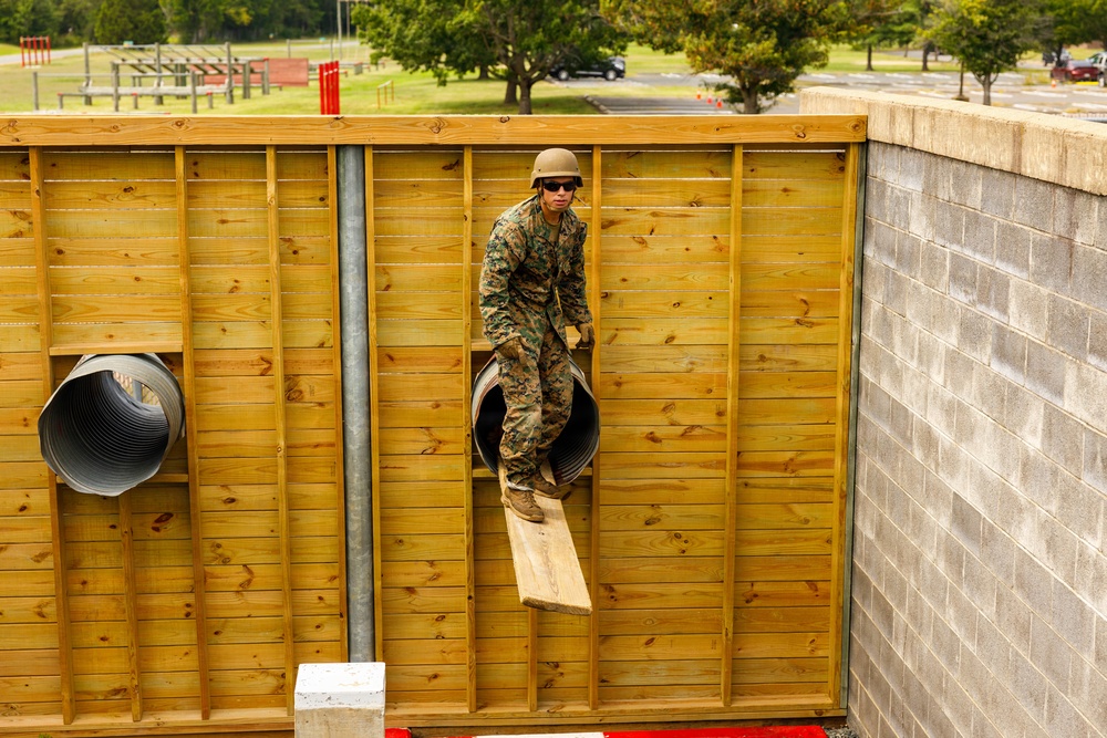 A military service member in camouflage uniform and helmet is navigating a wooden obstacle course wall, emerging through a cylindrical tunnel opening. The setting appears to be a military training facility with additional obstacle course elements visible in the background. The soldier is in the process of climbing out of or through a corrugated metal tube embedded in the wooden wall structure.