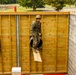 A military service member in camouflage uniform and helmet is navigating a wooden obstacle course wall, emerging through a cylindrical tunnel opening. The setting appears to be a military training facility with additional obstacle course elements visible in the background. The soldier is in the process of climbing out of or through a corrugated metal tube embedded in the wooden wall structure.