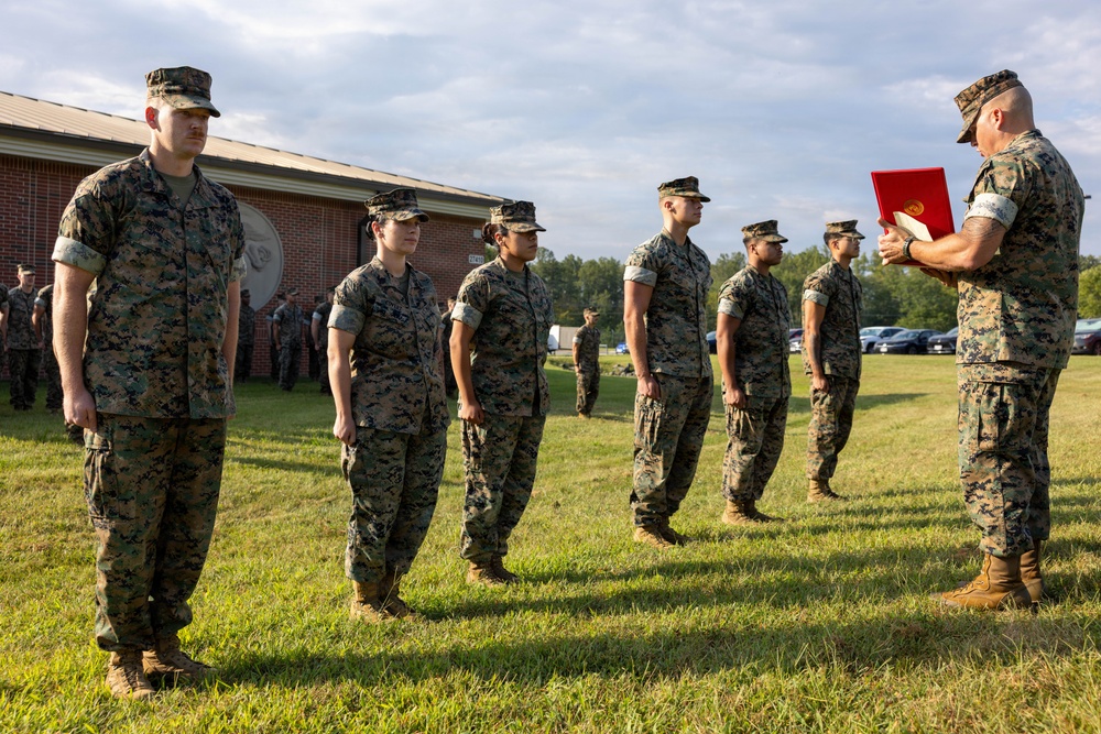 A military ceremony is taking place outdoors on a grass field, likely at a U.S. Marine Corps installation. A senior officer or NCO on the right is holding a red folder and appears to be reading from it, while a line of Marines in MARPAT camouflage uniforms stand at attention. This appears to be an awards, promotion, or recognition ceremony.