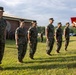 A military ceremony is taking place outdoors on a grass field, likely at a U.S. Marine Corps installation. A senior officer or NCO on the right is holding a red folder and appears to be reading from it, while a line of Marines in MARPAT camouflage uniforms stand at attention. This appears to be an awards, promotion, or recognition ceremony.