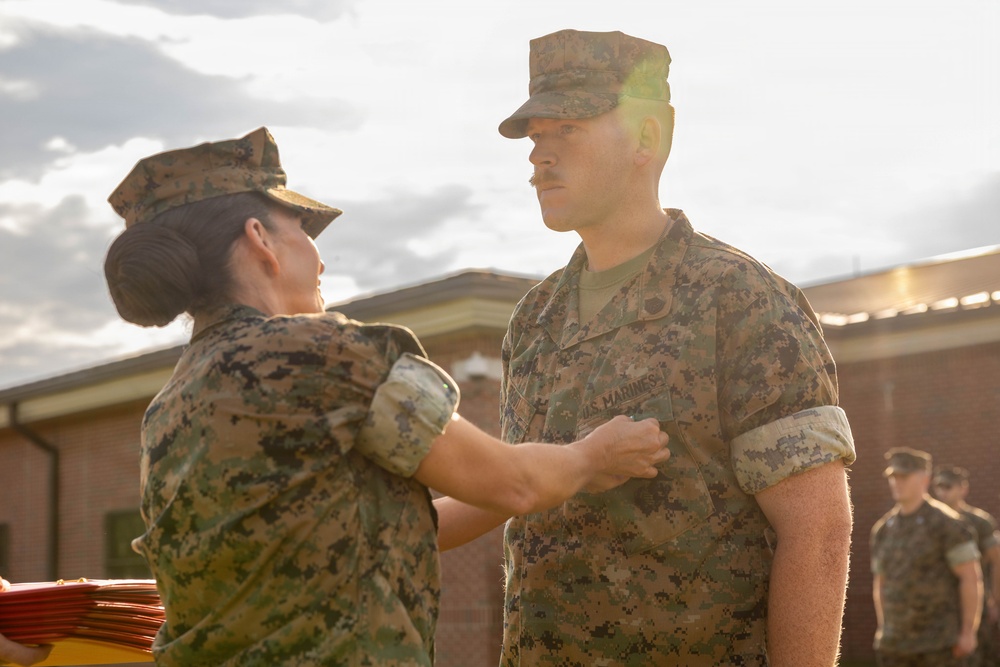 Two U.S. Marines in MARPAT camouflage uniforms are engaged in what appears to be a military awards or pinning ceremony outdoors. A female Marine is pinning something onto the chest of a male Marine who stands at attention. Additional Marines are visible in the background, and red folders or certificates are stacked nearby.