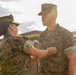 Two U.S. Marines in MARPAT camouflage uniforms are engaged in what appears to be a military awards or pinning ceremony outdoors. A female Marine is pinning something onto the chest of a male Marine who stands at attention. Additional Marines are visible in the background, and red folders or certificates are stacked nearby.