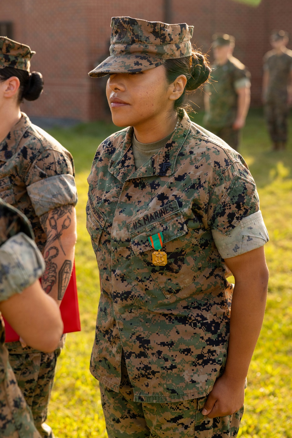 A U.S. Marine in MARPAT woodland camouflage uniform stands at attention outdoors, wearing a recently awarded medal on her chest. The medal appears to be a Navy and Marine Corps Achievement Medal, suspended from a green and orange ribbon. The setting appears to be a formal awards ceremony on a military installation.