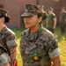 A U.S. Marine in MARPAT woodland camouflage uniform stands at attention outdoors, wearing a recently awarded medal on her chest. The medal appears to be a Navy and Marine Corps Achievement Medal, suspended from a green and orange ribbon. The setting appears to be a formal awards ceremony on a military installation.