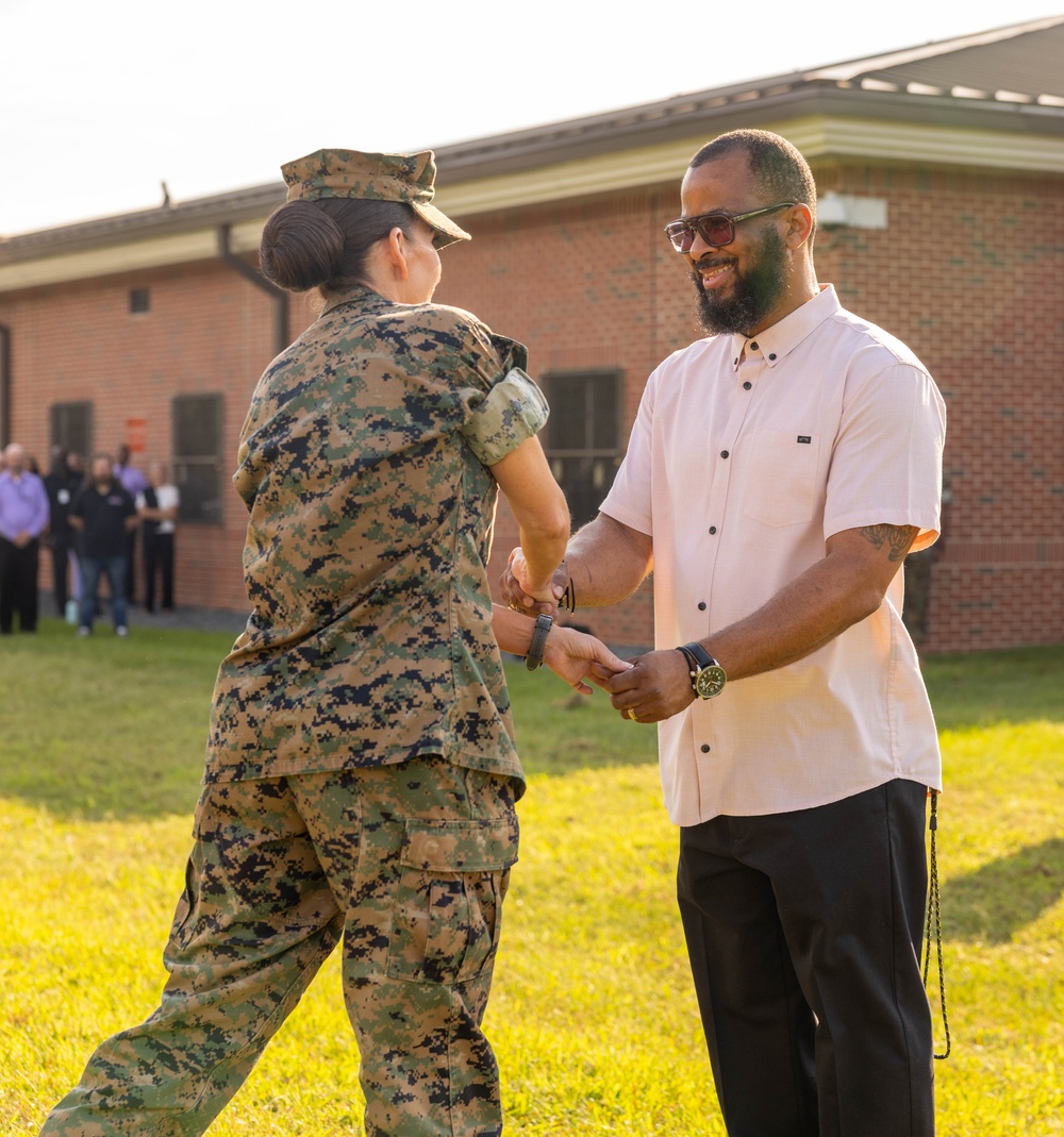 A U.S. Marine in MARPAT digital camouflage uniform is presenting or exchanging an item with a civilian man in a light-colored button-up shirt on a grassy field. The civilian is smiling and appears to be receiving something, suggesting a formal recognition or award ceremony. A group of observers is visible in the background near a brick building.