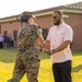 A U.S. Marine in MARPAT digital camouflage uniform is presenting or exchanging an item with a civilian man in a light-colored button-up shirt on a grassy field. The civilian is smiling and appears to be receiving something, suggesting a formal recognition or award ceremony. A group of observers is visible in the background near a brick building.