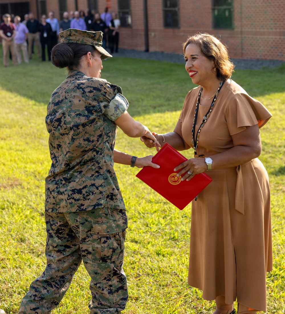 A female military service member in MARPAT digital camouflage uniform is presenting or exchanging a red official folder/certificate with a civilian woman in a tan dress during what appears to be a formal outdoor ceremony. The civilian woman is smiling and holding a red folder bearing a gold seal, suggesting an award, recognition, or official presentation. A group of observers is visible in the background near a brick building.