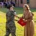 A female military service member in MARPAT digital camouflage uniform is presenting or exchanging a red official folder/certificate with a civilian woman in a tan dress during what appears to be a formal outdoor ceremony. The civilian woman is smiling and holding a red folder bearing a gold seal, suggesting an award, recognition, or official presentation. A group of observers is visible in the background near a brick building.