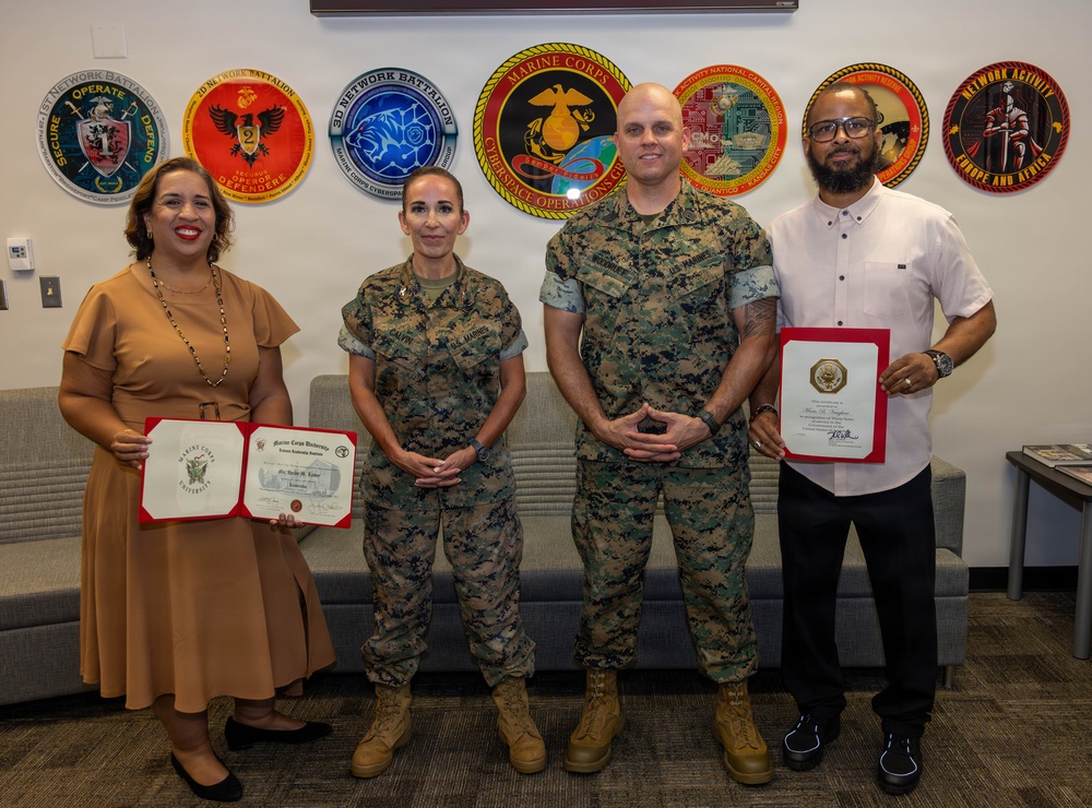 Four individuals pose for a formal photo in what appears to be a U.S. Marine Corps facility. Two are in Marine Corps Combat Utility Uniforms (MCCUU) and two are in civilian attire, with the civilians holding red-covered certificates or awards. The wall behind them displays multiple unit insignia patches from various Marine Corps Network Battalions and Cyberspace Operations units.