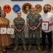 Four individuals pose for a formal photo in what appears to be a U.S. Marine Corps facility. Two are in Marine Corps Combat Utility Uniforms (MCCUU) and two are in civilian attire, with the civilians holding red-covered certificates or awards. The wall behind them displays multiple unit insignia patches from various Marine Corps Network Battalions and Cyberspace Operations units.