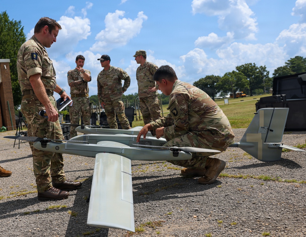 Military personnel in camouflage uniforms are gathered around a fixed-wing unmanned aerial vehicle (UAV) on a gravel surface outdoors. One soldier is crouching and inspecting or adjusting the drone while others observe, suggesting a pre-flight check, training exercise, or equipment demonstration. The setting appears to be a military installation or training area.