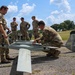 Military personnel in camouflage uniforms are gathered around a fixed-wing unmanned aerial vehicle (UAV) on a gravel surface outdoors. One soldier is crouching and inspecting or adjusting the drone while others observe, suggesting a pre-flight check, training exercise, or equipment demonstration. The setting appears to be a military installation or training area.