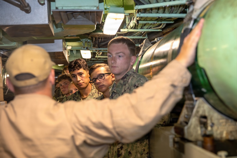 An instructor or senior personnel wearing a tan uniform and cap is briefing a group of young military personnel in digital camouflage uniforms inside what appears to be a submarine or naval vessel. The instructor is pointing toward a large cylindrical green object, likely a torpedo or missile tube. The group of trainees is attentively observing the demonstration in a confined, equipment-filled space.