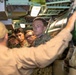 An instructor or senior personnel wearing a tan uniform and cap is briefing a group of young military personnel in digital camouflage uniforms inside what appears to be a submarine or naval vessel. The instructor is pointing toward a large cylindrical green object, likely a torpedo or missile tube. The group of trainees is attentively observing the demonstration in a confined, equipment-filled space.