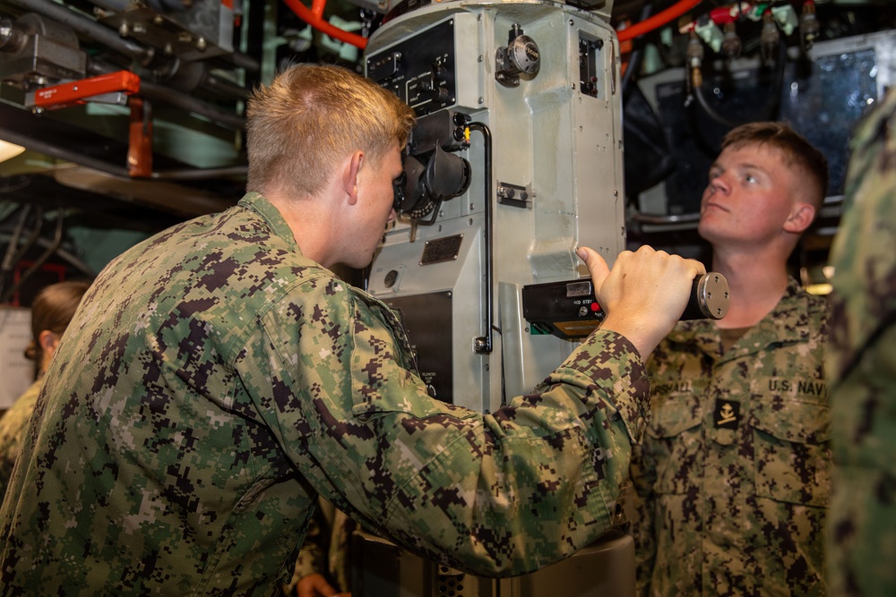 Two U.S. Navy sailors in NWU Type III (woodland camouflage) uniforms are working with a periscope or optical instrument inside what appears to be a submarine. One sailor is looking through the eyepiece while the other assists by holding a flashlight and observing. The confined space with overhead pipes, cables, and equipment is consistent with a submarine interior.