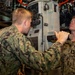 Two U.S. Navy sailors in NWU Type III (woodland camouflage) uniforms are working with a periscope or optical instrument inside what appears to be a submarine. One sailor is looking through the eyepiece while the other assists by holding a flashlight and observing. The confined space with overhead pipes, cables, and equipment is consistent with a submarine interior.