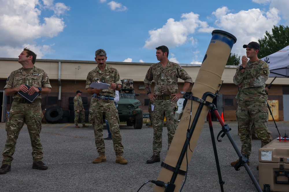 A group of military personnel in multicam/OCP camouflage uniforms are gathered in an outdoor area, apparently during a briefing or equipment demonstration. A mortar tube mounted on a bipod stand is prominently visible in the foreground. The setting appears to be a military installation or training area with vehicles and a building visible in the background.