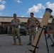 A group of military personnel in multicam/OCP camouflage uniforms are gathered in an outdoor area, apparently during a briefing or equipment demonstration. A mortar tube mounted on a bipod stand is prominently visible in the foreground. The setting appears to be a military installation or training area with vehicles and a building visible in the background.