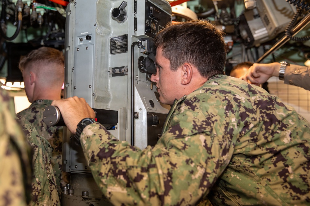 Military personnel in camouflage uniforms are operating or inspecting equipment inside what appears to be a naval vessel or military vehicle. The central figure is looking through an optical viewing device while another service member assists with controls. The setting appears to be a confined interior space with complex instrumentation and machinery.