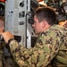 Military personnel in camouflage uniforms are operating or inspecting equipment inside what appears to be a naval vessel or military vehicle. The central figure is looking through an optical viewing device while another service member assists with controls. The setting appears to be a confined interior space with complex instrumentation and machinery.
