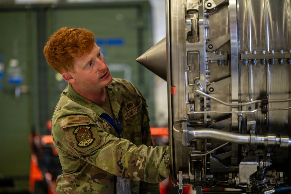A U.S. Air Force airman in OCP (Operational Camouflage Pattern) uniform is working on what appears to be a jet engine or turbine component in an indoor maintenance facility. The individual is closely inspecting or servicing the mechanical assembly, which features visible fuel lines, casing components, and a conical center body typical of jet engine inlets or afterburner sections. The setting appears to be a military aircraft maintenance hangar.