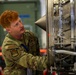 A U.S. Air Force airman in OCP (Operational Camouflage Pattern) uniform is working on what appears to be a jet engine or turbine component in an indoor maintenance facility. The individual is closely inspecting or servicing the mechanical assembly, which features visible fuel lines, casing components, and a conical center body typical of jet engine inlets or afterburner sections. The setting appears to be a military aircraft maintenance hangar.