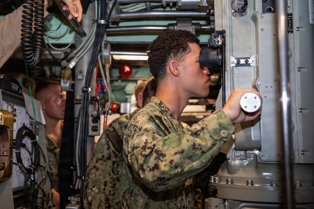 A U.S. Navy sailor in NWU Type III (woodland digital camouflage) uniform is looking through an optical viewing device, likely a periscope or targeting optic, aboard what appears to be a naval vessel or submarine. The sailor is operating controls with one hand while peering through the eyepiece. Additional personnel in similar uniforms are visible in the background within the confined, equipment-dense space.
