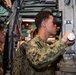 A U.S. Navy sailor in NWU Type III (woodland digital camouflage) uniform is looking through an optical viewing device, likely a periscope or targeting optic, aboard what appears to be a naval vessel or submarine. The sailor is operating controls with one hand while peering through the eyepiece. Additional personnel in similar uniforms are visible in the background within the confined, equipment-dense space.