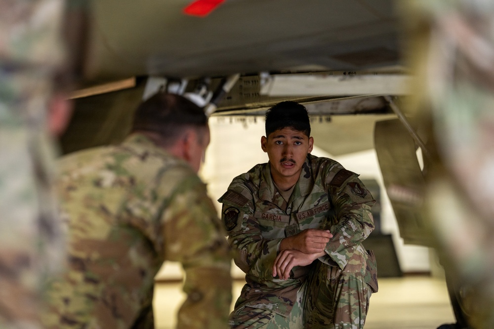 A U.S. Air Force servicemember named Garcia is crouching beneath what appears to be a military aircraft, engaged in conversation with another servicemember whose back is to the camera. Both individuals are wearing OCP (Operational Camouflage Pattern) uniforms. The setting appears to be a flight line or hangar environment.