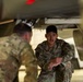 A U.S. Air Force servicemember named Garcia is crouching beneath what appears to be a military aircraft, engaged in conversation with another servicemember whose back is to the camera. Both individuals are wearing OCP (Operational Camouflage Pattern) uniforms. The setting appears to be a flight line or hangar environment.