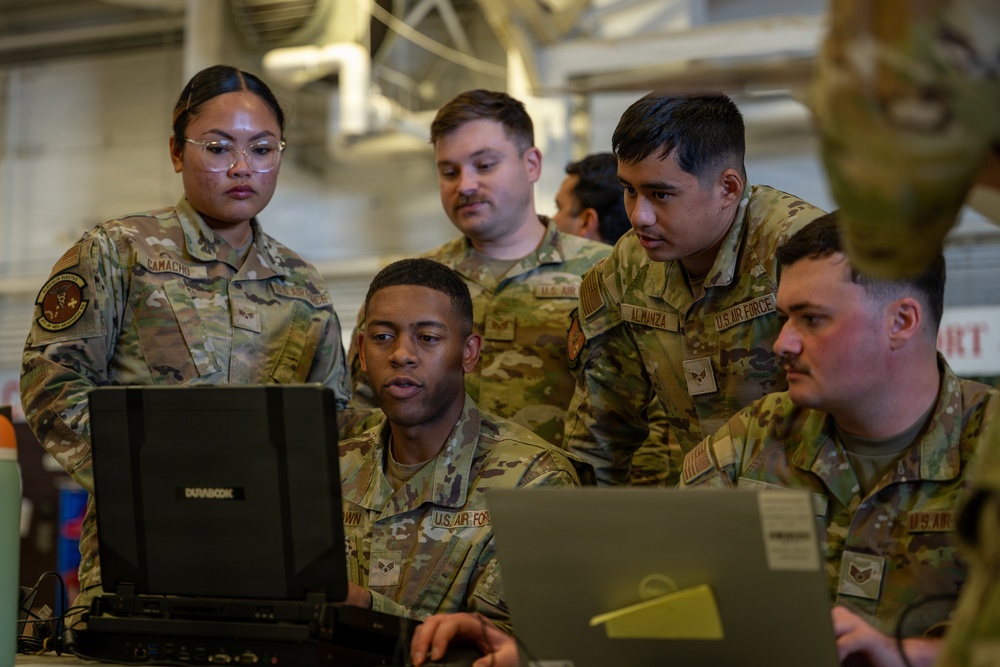 A group of U.S. Air Force personnel in OCP (Operational Camouflage Pattern) uniforms are gathered around laptop computers in what appears to be a hangar or maintenance facility. Several airmen are focused on a Durabook ruggedized laptop, with one airman appearing to brief or explain something on the screen. Name tapes visible include 'CAMACHO' and 'ALMANZA', and 'U.S. AIR FORCE' tape is visible on multiple uniforms.