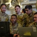 A group of U.S. Air Force personnel in OCP (Operational Camouflage Pattern) uniforms are gathered around laptop computers in what appears to be a hangar or maintenance facility. Several airmen are focused on a Durabook ruggedized laptop, with one airman appearing to brief or explain something on the screen. Name tapes visible include 'CAMACHO' and 'ALMANZA', and 'U.S. AIR FORCE' tape is visible on multiple uniforms.