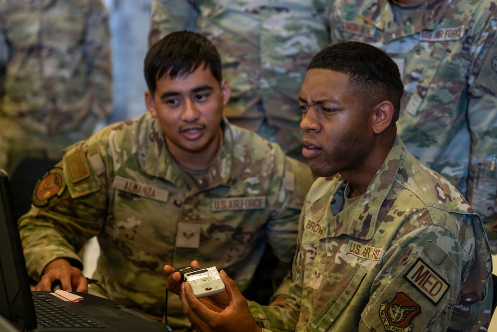 Two U.S. Air Force personnel in OCP (Operational Camouflage Pattern) uniforms are working together at a laptop computer. The airman in the foreground, identified by nametape as 'BROWN' with a 'MED' patch, is holding a small device while discussing something with 'ALMANZA' beside him. Additional uniformed personnel are visible in the background.