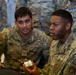 Two U.S. Air Force personnel in OCP (Operational Camouflage Pattern) uniforms are working together at a laptop computer. The airman in the foreground, identified by nametape as 'BROWN' with a 'MED' patch, is holding a small device while discussing something with 'ALMANZA' beside him. Additional uniformed personnel are visible in the background.