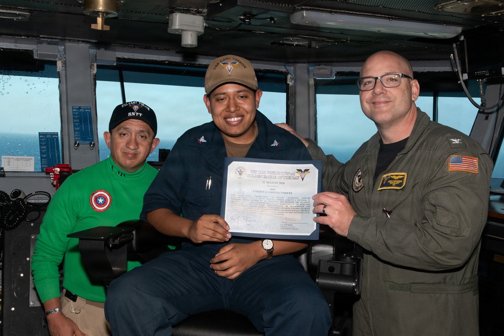 Three individuals pose for a photo aboard what appears to be the bridge of USS Carl Vinson (CVN-70), a U.S. Navy aircraft carrier. The central figure is holding a certificate that reads 'USS Carl Vinson Golden Eagle of the Day' dated 22 August 2026, awarded to 'Enrique J. Cabrera-Vasquez.' The occasion appears to be a recognition ceremony for the sailor in the center.
