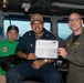 Three individuals pose for a photo aboard what appears to be the bridge of USS Carl Vinson (CVN-70), a U.S. Navy aircraft carrier. The central figure is holding a certificate that reads 'USS Carl Vinson Golden Eagle of the Day' dated 22 August 2026, awarded to 'Enrique J. Cabrera-Vasquez.' The occasion appears to be a recognition ceremony for the sailor in the center.